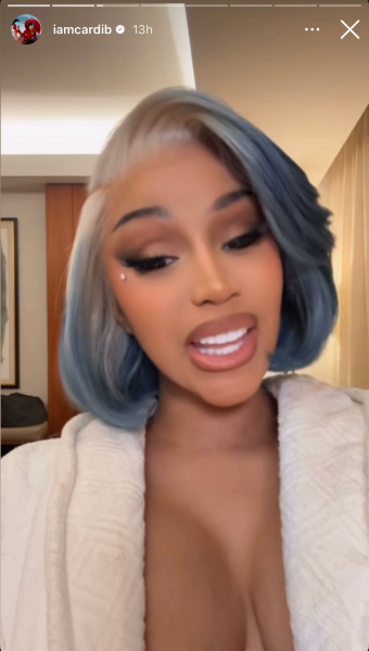 Cardi B talks to the camera. She wears a robe and her hair is styled in a blue bob with platinum blonde roots.